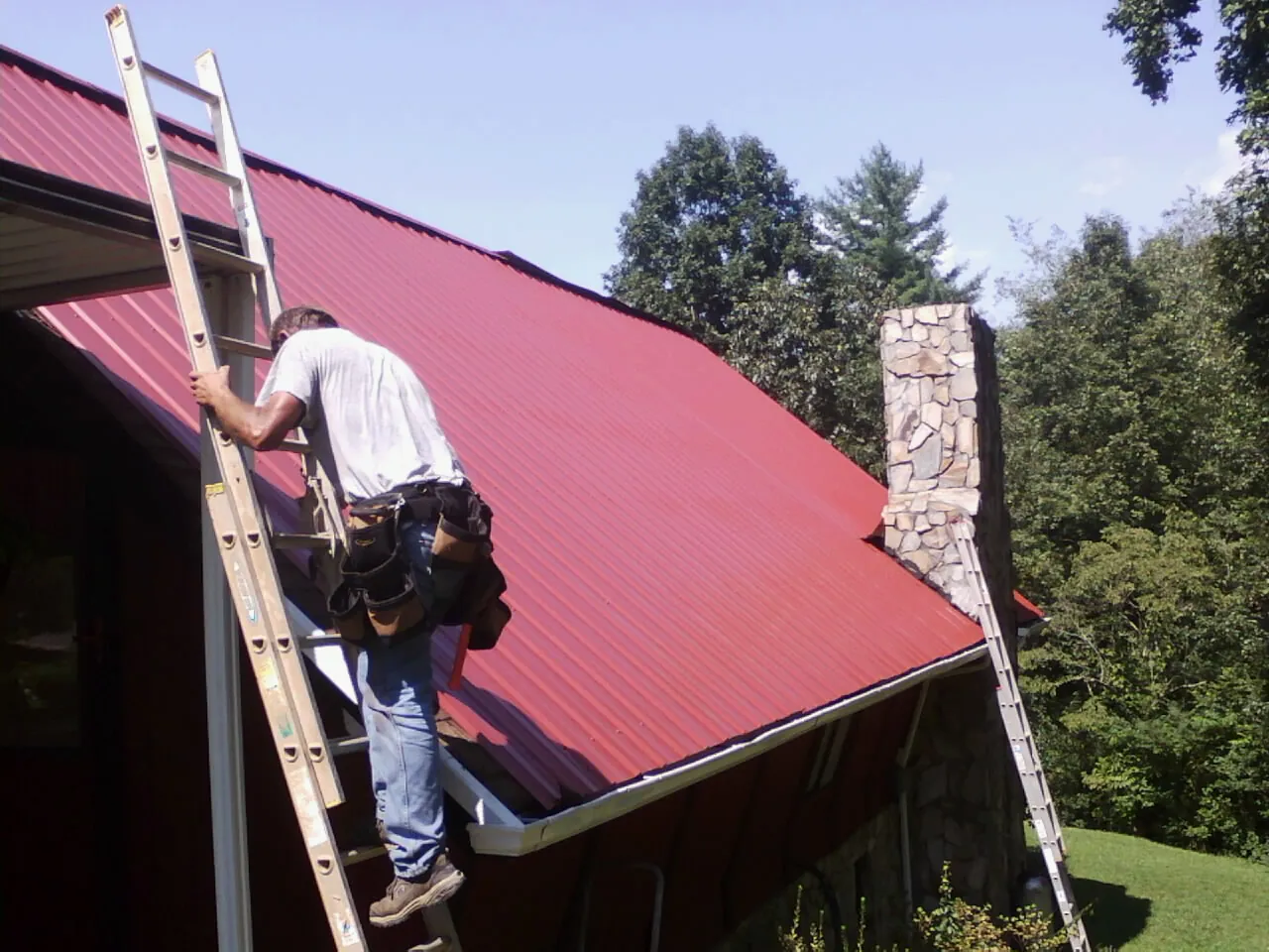 Roofer on a ladder beside a red metal roof and stone chimney — Soffit & Fascia Repair by Roofing Brothers in Shippingport